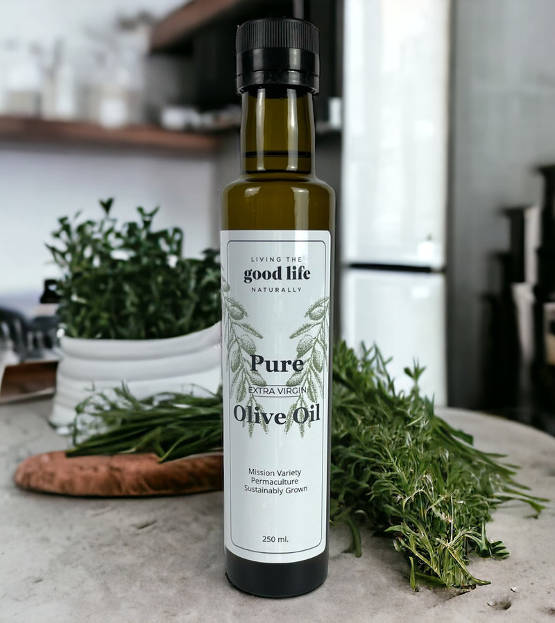 A dark green narrow bottle with a white label. The bottle sits on a kitchen counter with herbs behind it. The label reads, "Living the Good Life Naturally, Pure Extra Virgin Olive Oil, Mission Variety, Permaculture, Sustainably Grown, 250mL."